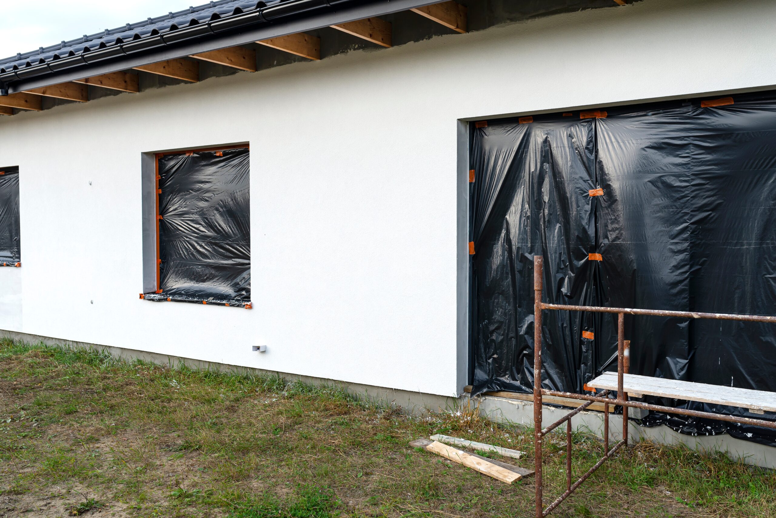 A newly applied layer of white silicone plaster on the wall of the house, visible metal scaffolding and boards, window is secured with black foil.
